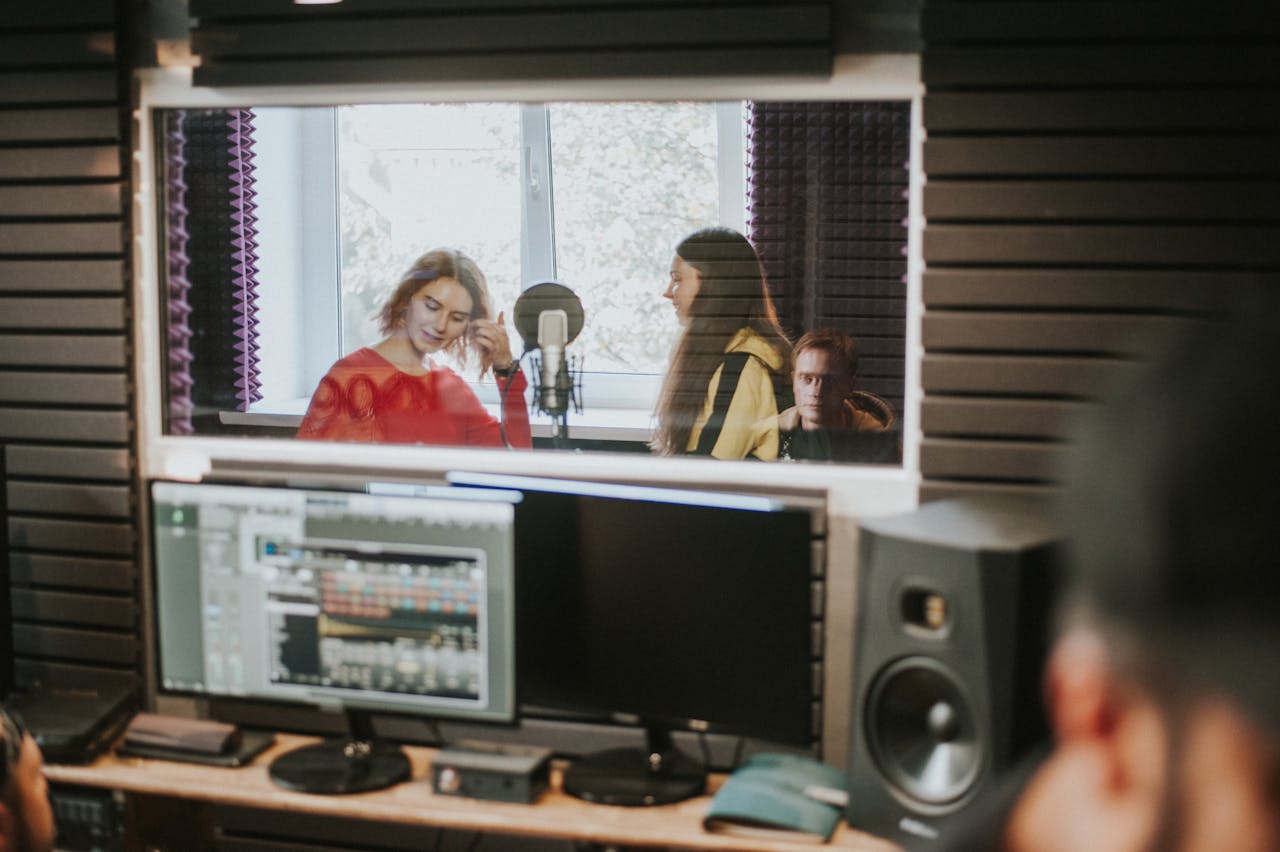Artists recording music in a professional studio setting, captured through a glass window.