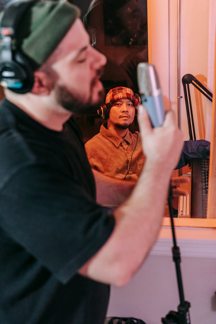 Two men collaborate in a recording studio. One sings into a microphone while the other observes.